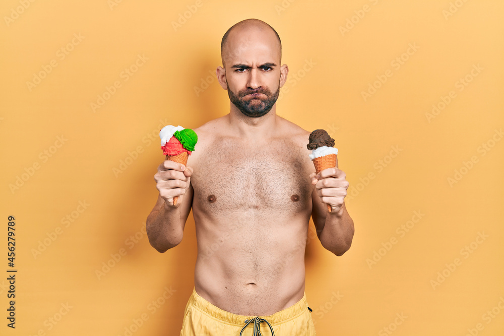 Fototapeta premium Young bald man wearing swimwear holding two ice cream cones skeptic and nervous, frowning upset because of problem. negative person.
