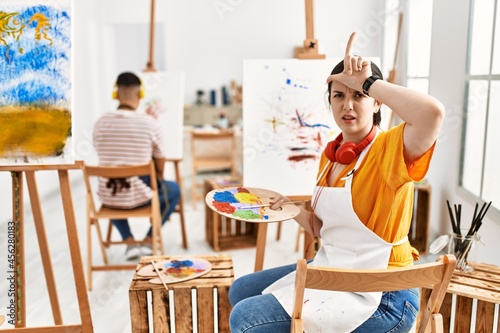Young artist woman painting on canvas at art studio making fun of people with fingers on forehead doing loser gesture mocking and insulting. © Krakenimages.com