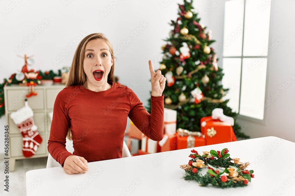 Caucasian young blonde woman sitting on the table by christmas tree pointing finger up with successful idea. exited and happy. number one. Caucasian young blonde woman sitting on the table by christmas tree pointing finger up with successful idea. exited and happy. number one.