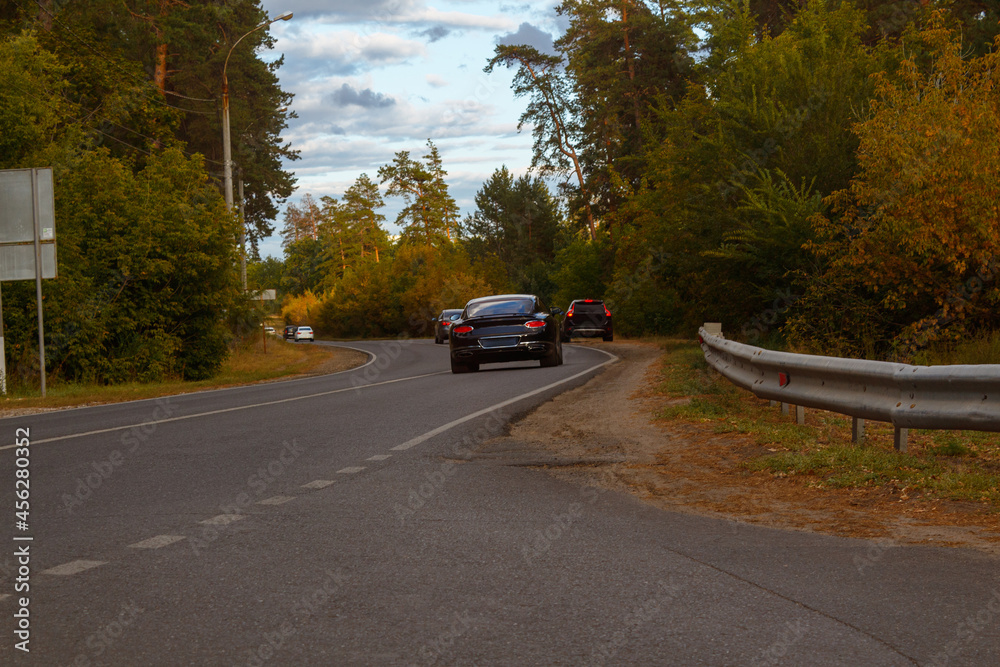 Autumn forest landscape. Asphalt road along the forest with a parked car on the side of the road. Cars drives along a forest road in autumn on a sunny day.