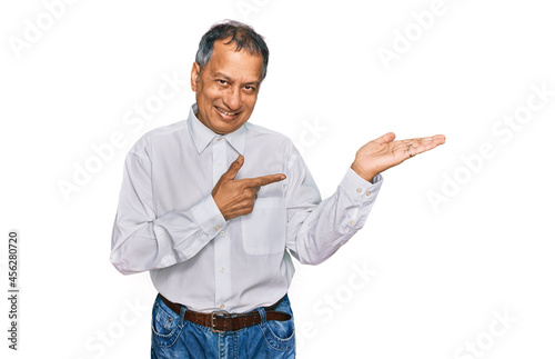 Middle age indian man wearing casual white shirt amazed and smiling to the camera while presenting with hand and pointing with finger.