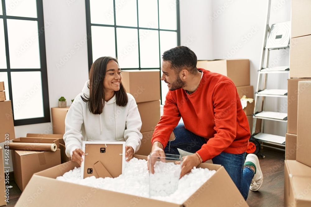 Young latin couple smiling happy unboxing cardboard box at new home.