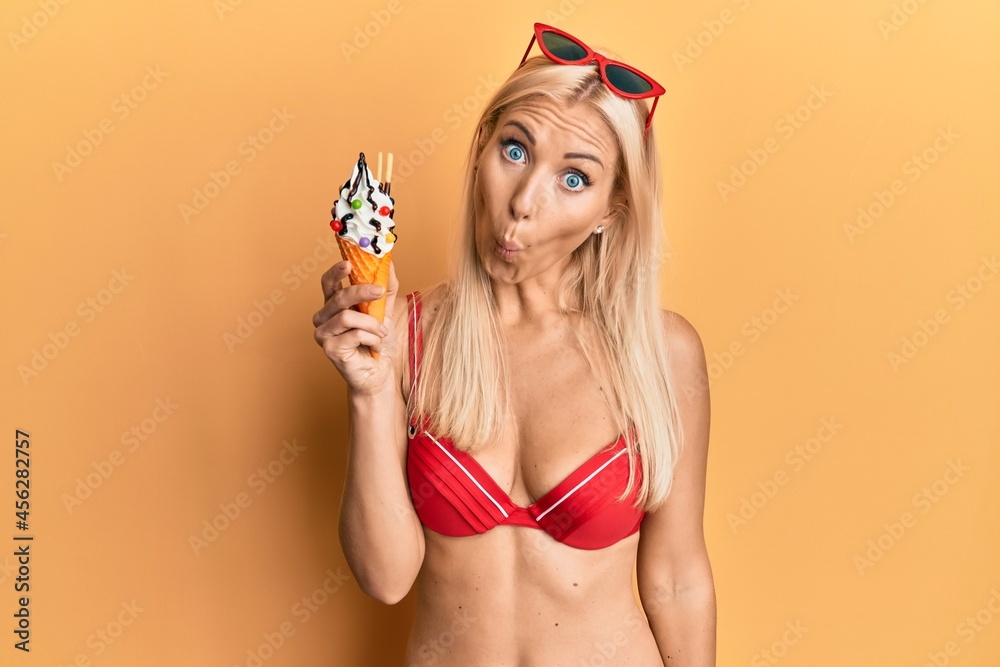 Young blonde woman wearing bikini holding ice cream scared and amazed with open mouth for surprise, disbelief face Young blonde woman wearing bikini holding ice cream scared and amazed with open mouth for surprise, disbelief face