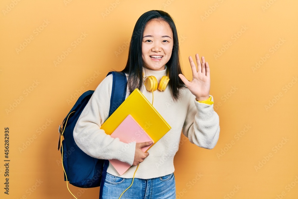 Young chinese girl holding student backpack and books waiving saying hello happy and smiling, friendly welcome gesture Young chinese girl holding student backpack and books waiving saying hello happy and smiling, friendly welcome gesture