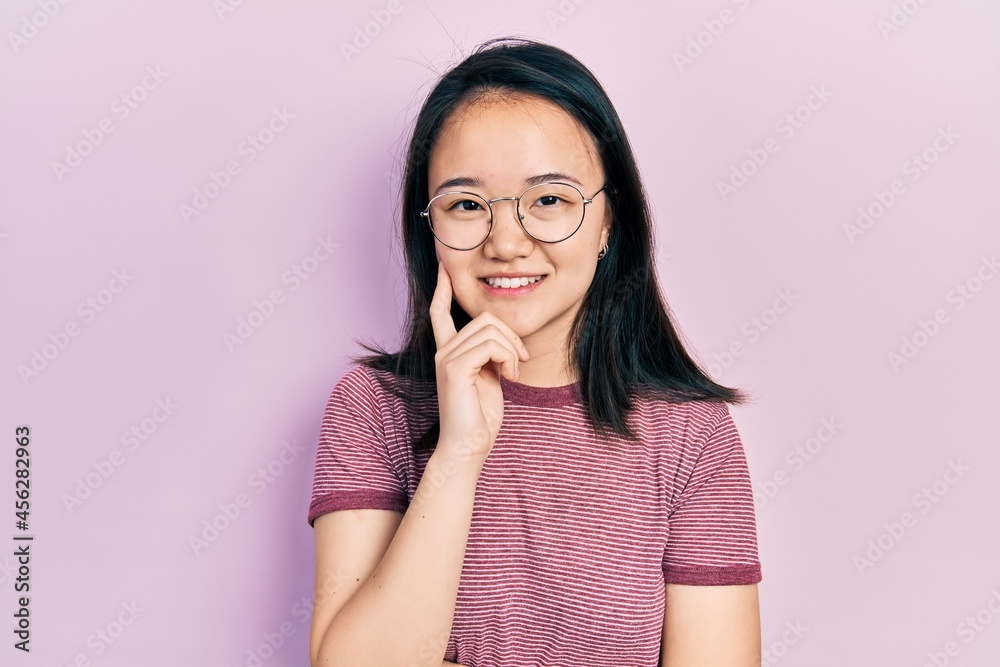 Young chinese girl wearing casual clothes and glasses looking confident at the camera with smile with crossed arms and hand raised on chin. thinking positive.