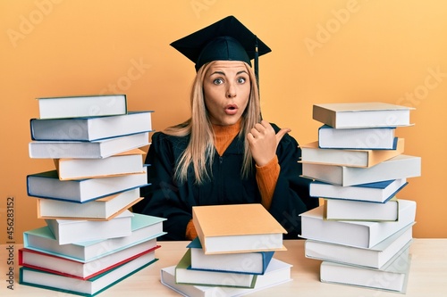 Young caucasian woman wearing graduation ceremony robe sitting on the table surprised pointing with hand finger to the side, open mouth amazed expression.