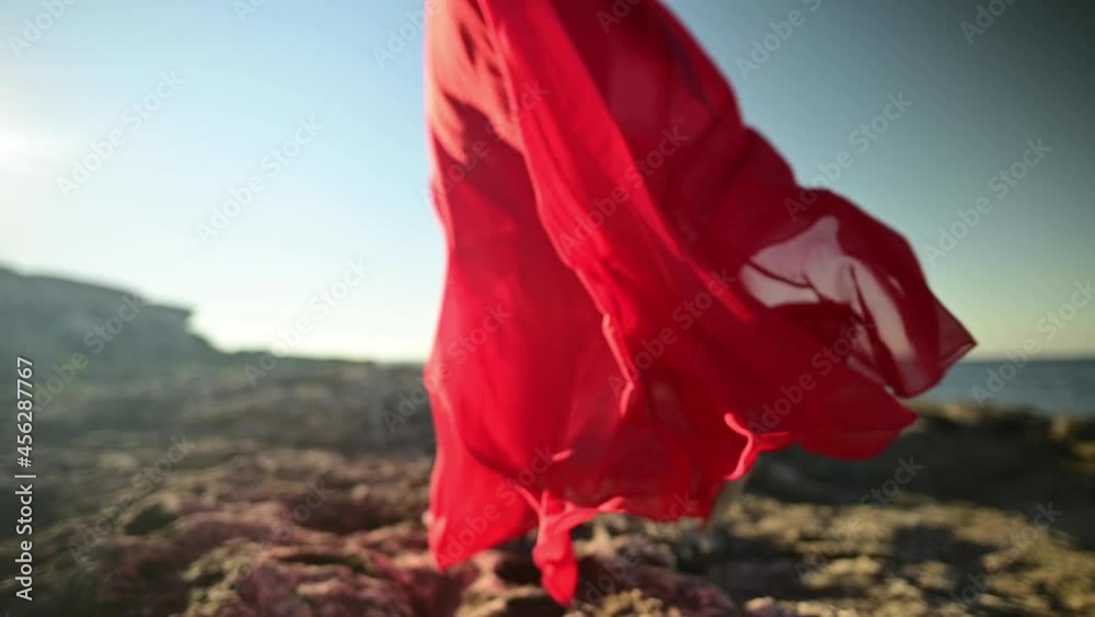 Slow motion young caucasian woman in a loose fluttering red dress is walking along the rocky seashore. Shallow depth of field. Offset focus.