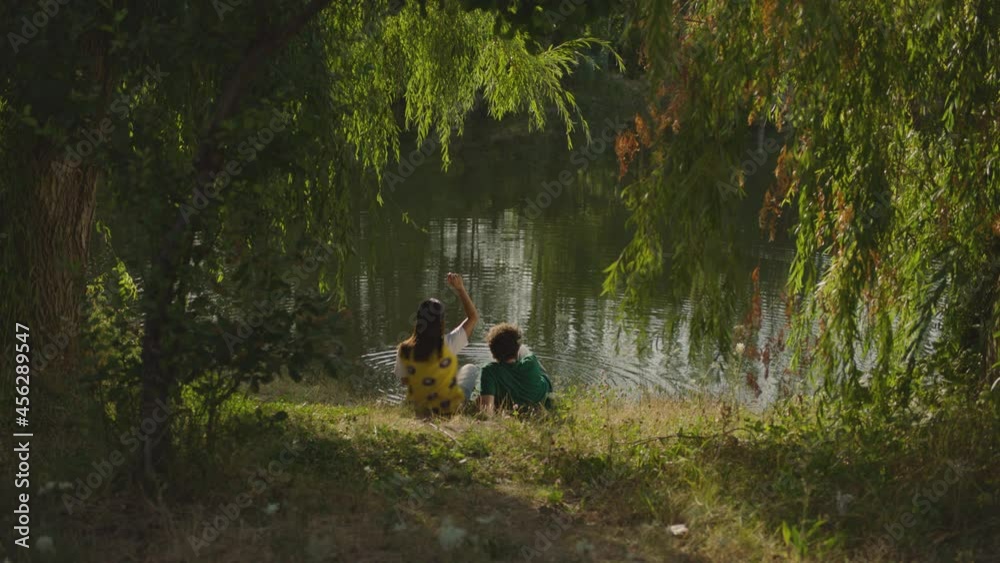 Young and Beautiful couple sitting under the tree by the lake inside park or forest . Boyfriend and girlfriend throwing stones into water and having fun . Back rear view . Slow Motion Young and Beautiful couple sitting under the tree by the lake inside park or forest . Boyfriend and girlfriend throwing stones into water and having fun . Back rear view . Slow Motion