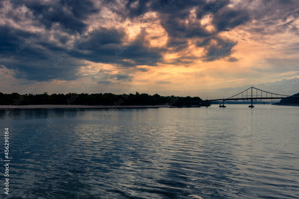 A wide river and a bridge under the setting sun behind the clouds. A calm river in the evening A wide river and a bridge under the setting sun behind the clouds. A calm river in the evening