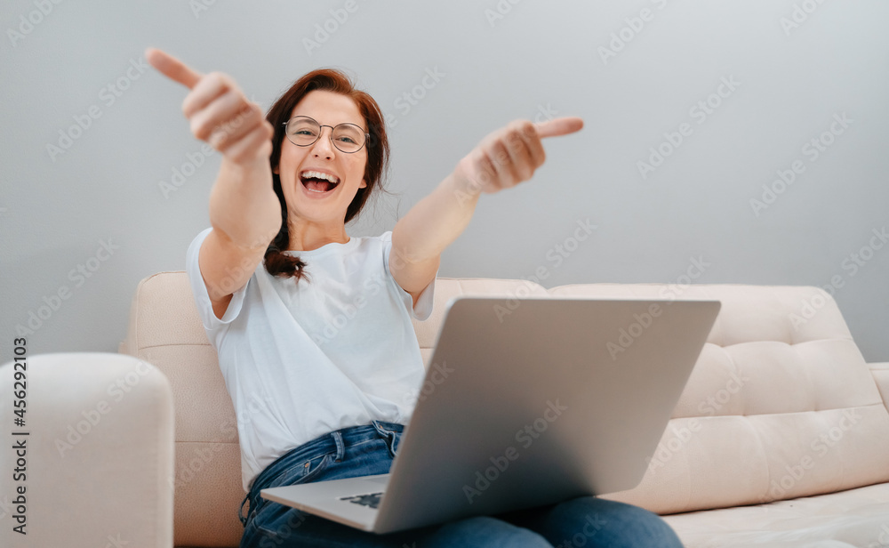 Smiling woman gives a thumbs-up to the camera while sitting with a computer on the couch at home. Concept of a satisfied user. Smiling woman gives a thumbs-up to the camera while sitting with a computer on the couch at home. Concept of a satisfied user.