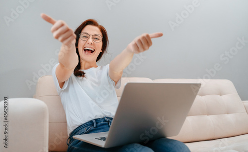 Smiling woman gives a thumbs-up to the camera while sitting with a computer on the couch at home. Concept of a satisfied user. © vadimverenitsyn