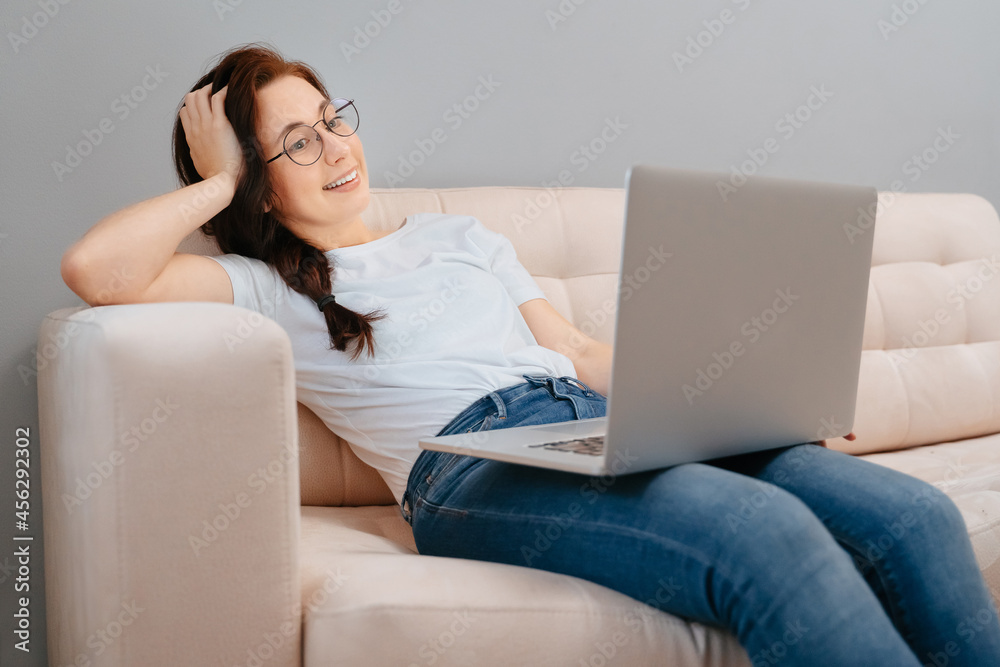 Naklejka premium Young happy woman with glasses is resting at home on the couch ordering goods online on a laptop. Concept of purchases and sales via the Internet.