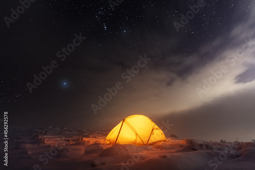 Yellow tent lighted from the inside against the backdrop of glowing city lights and incredible starry sky. Amazing night landscape. Tourists camp in winter mountains. Travel concept