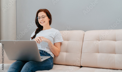 Young satisfied woman sitting on the sofa in the living room of the house listens to the interlocutor on the laptop online.