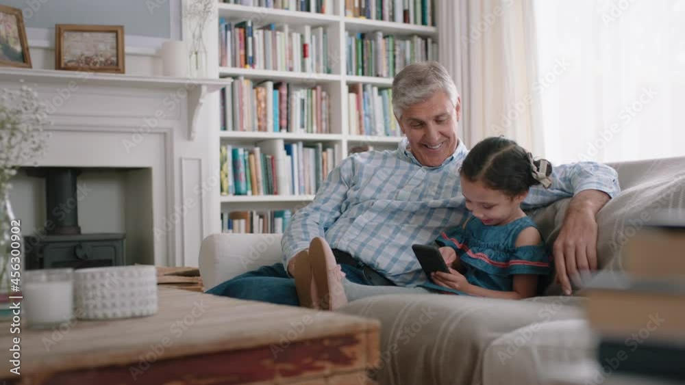 happy grandfather showing little girl how to use smartphone teaching curious granddaughter modern technology intelligent child learning mobile phone sitting with grandpa on sofa