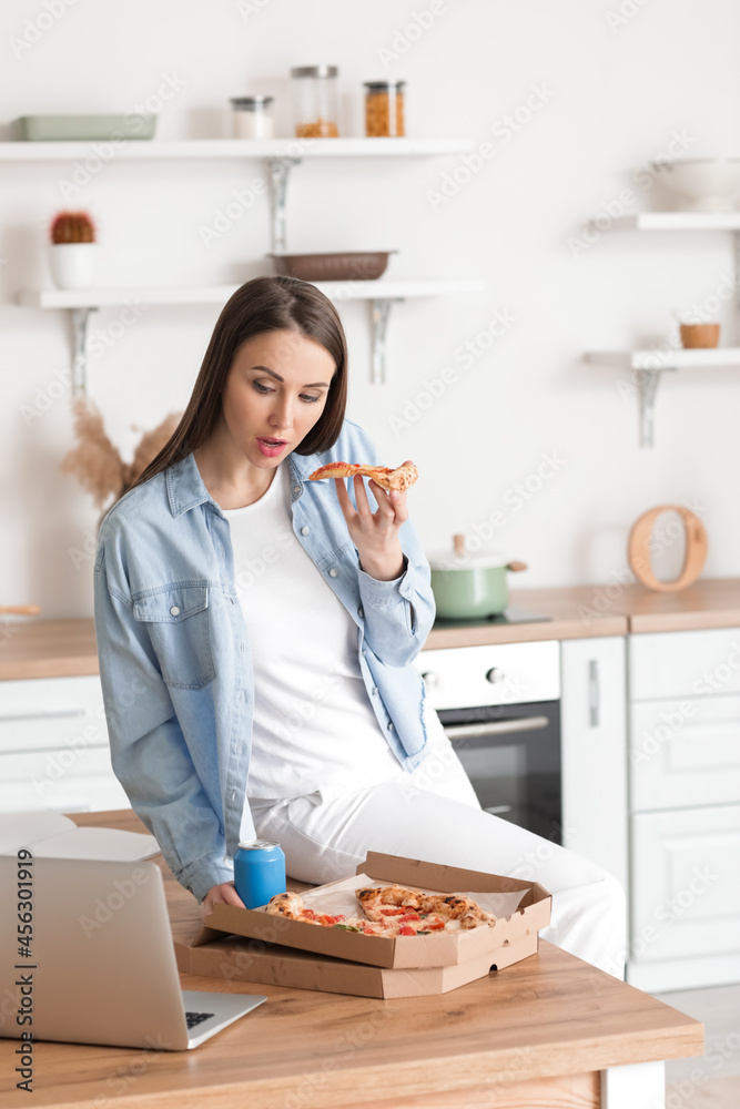 Beautiful young woman with laptop eating tasty pizza in kitchen Beautiful young woman with laptop eating tasty pizza in kitchen