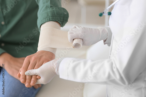 Doctor applying bandage onto arm of young man in clinic, closeup