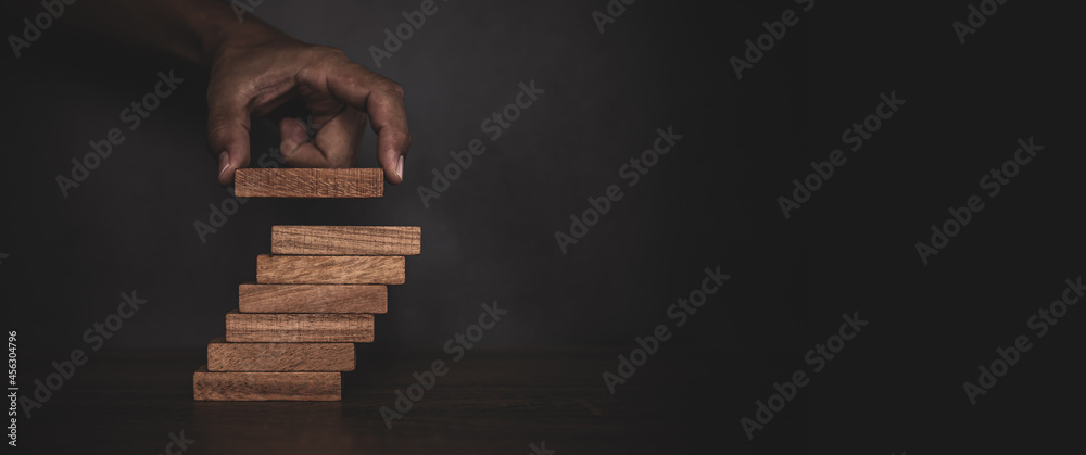 Close-up hand is placing wood block tower stacked in stair step with caution to prevent collapse or crash concepts of financial risk management and strategic planning. - obrazy, fototapety, plakaty 