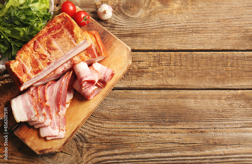 Board with tasty smoked bacon and fresh vegetables on wooden background