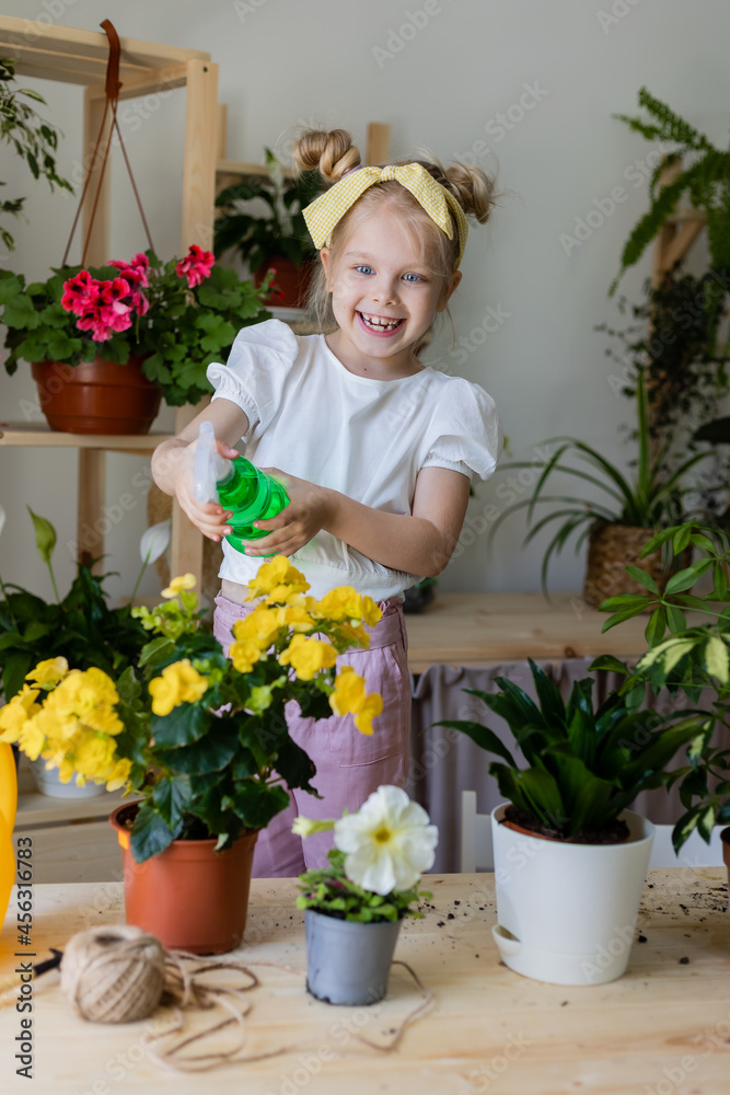 a little blonde girl with a headband on her head and wearing work gloves sprays from a bullet dispenser or waters indoor flowers. Home gardening, caring houseplants. Space for text. High quality photo a little blonde girl with a headband on her head and wearing work gloves sprays from a bullet dispenser or waters indoor flowers. Home gardening, caring houseplants. Space for text. High quality photo