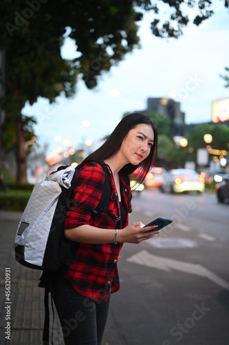 Young Asian woman standing near the road with a smartphone in her hand and waiting for a taxi.