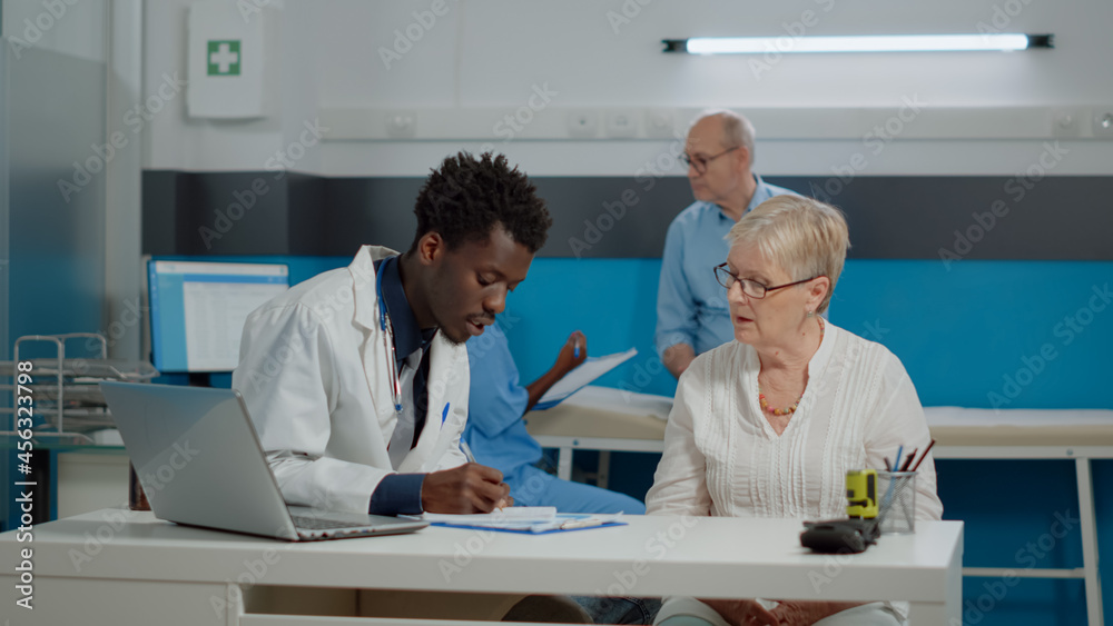 Caucasian aged patient receiving consultation from doctor in medical office at facility. Medic examining disease symptoms for sick older woman while nurse discussing with senior man Caucasian aged patient receiving consultation from doctor in medical office at facility. Medic examining disease symptoms for sick older woman while nurse discussing with senior man