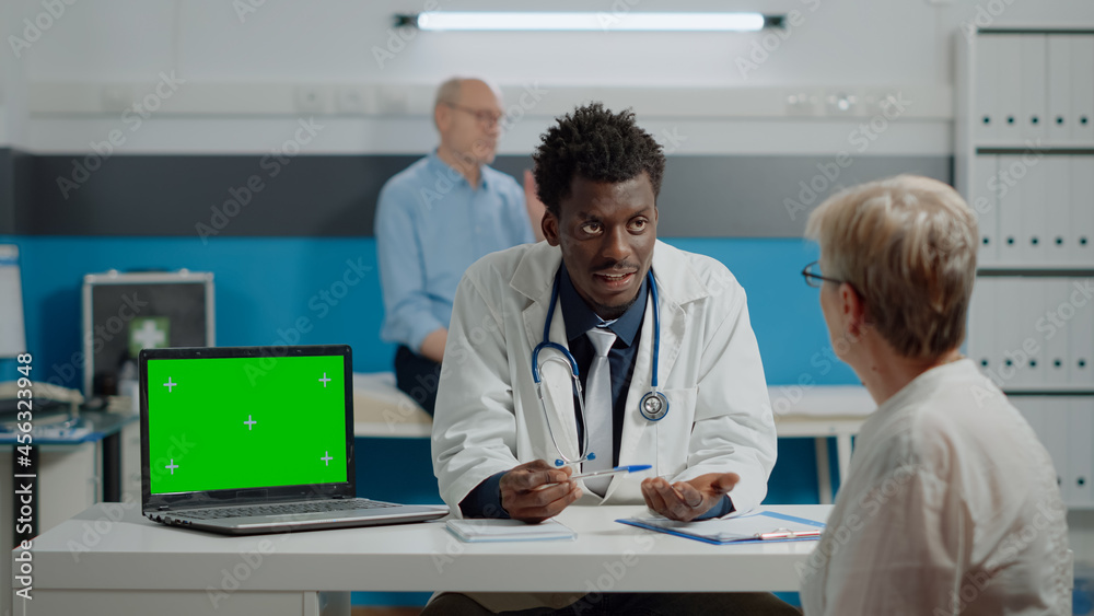 Adult specialist talking to patient with horizontal green screen on laptop in medical office. Young medic having device with chroma key isolated background and mockup template on desk