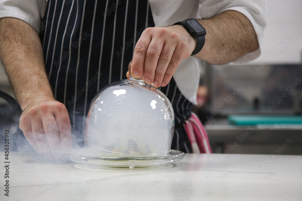 Chef's hand lifts up glass cloche from a plate with hot food and moving smoke at the restaurant. Exquisite dish, creative restaurant meal concept, haute couture food.