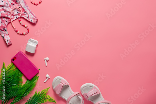 Summer top view concept. Girl's swimsuit and sandals with accessories on the pink background with green leaves. 