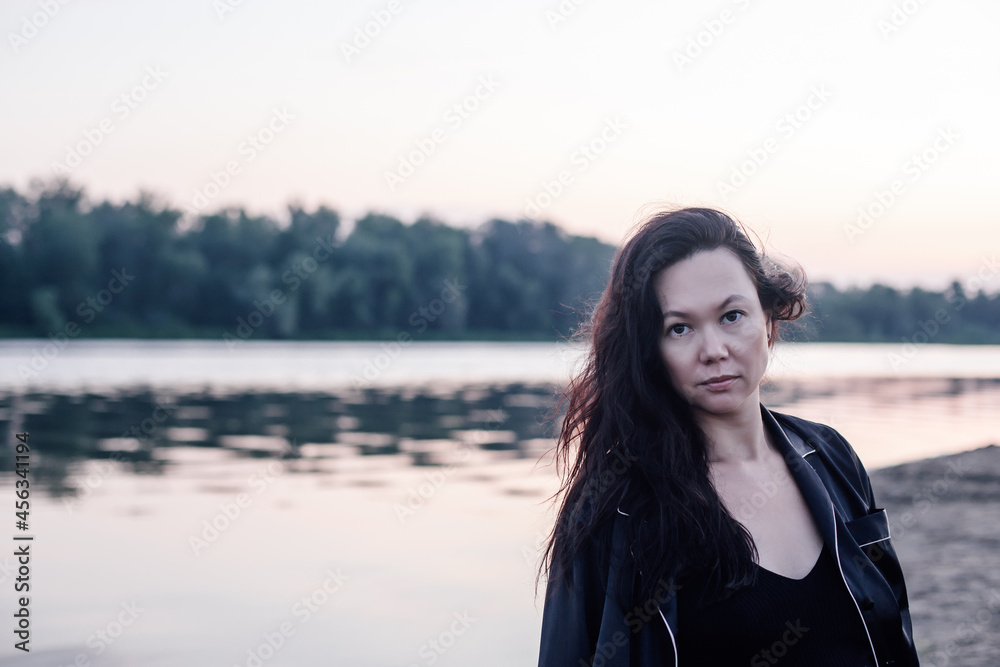 Portrait of woman against background of forest and pond. Young attractive woman in close-up in dark jacket looks at camera and stands on shore of picturesque lake. Portrait of woman against background of forest and pond. Young attractive woman in close-up in dark jacket looks at camera and stands on shore of picturesque lake.