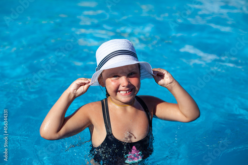 Child in white panama is enjoying summer. Little happy tanned girl in black swimsuit stands in water and holds white straw hat decorated with black ribbon in her hands and laughs. 