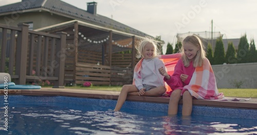 Portrait of happy sisters wrapped in towel sitting near swimming pool