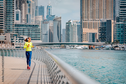 Happy woman wearing headphones and sunglasses trains and does running and fitness on the sidewalk of the embankment in the Dubai Marina area