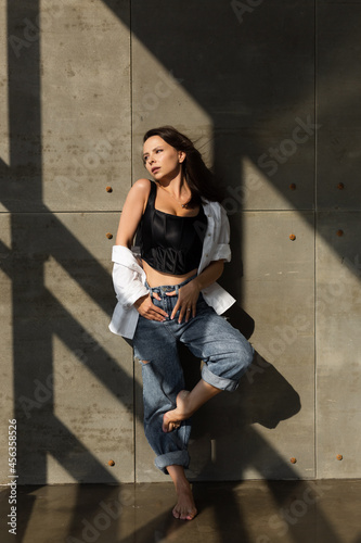 Pretty young woman with big breast posing in studio, by steel wall on background, wearing jeans and white shirt, natural warm sunset light