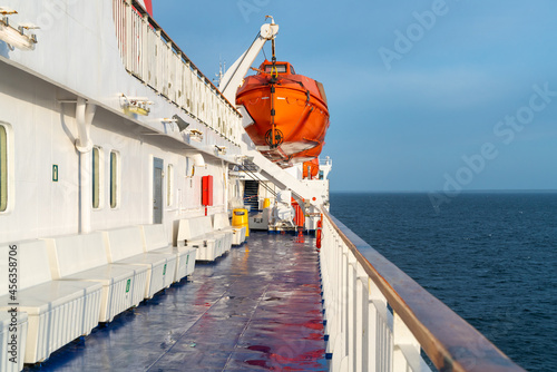 Orange life boat hanging on a crane on a deck of sailing ocean ship with ocean horizon in the back and wooden railing in front