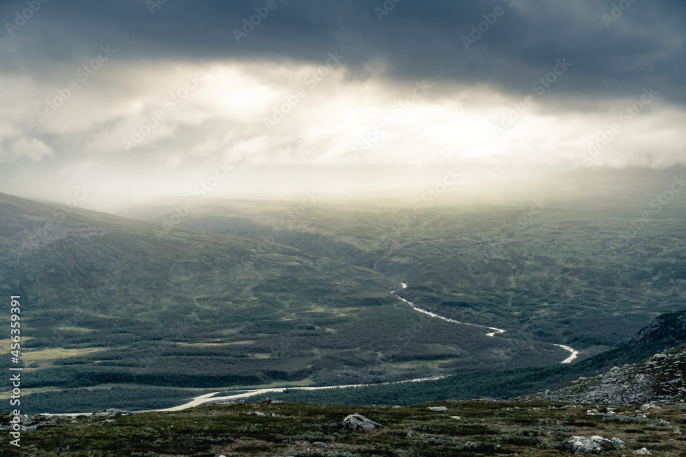 Bright sunlight comes through thick clouds and illuminates heavy rainfall in the distance above remote rough arctic valley. Dramatic weather in Rapadalen, Sarek National park, Swedish Lapland. Bright sunlight comes through thick clouds and illuminates heavy rainfall in the distance above remote rough arctic valley. Dramatic weather in Rapadalen, Sarek National park, Swedish Lapland.