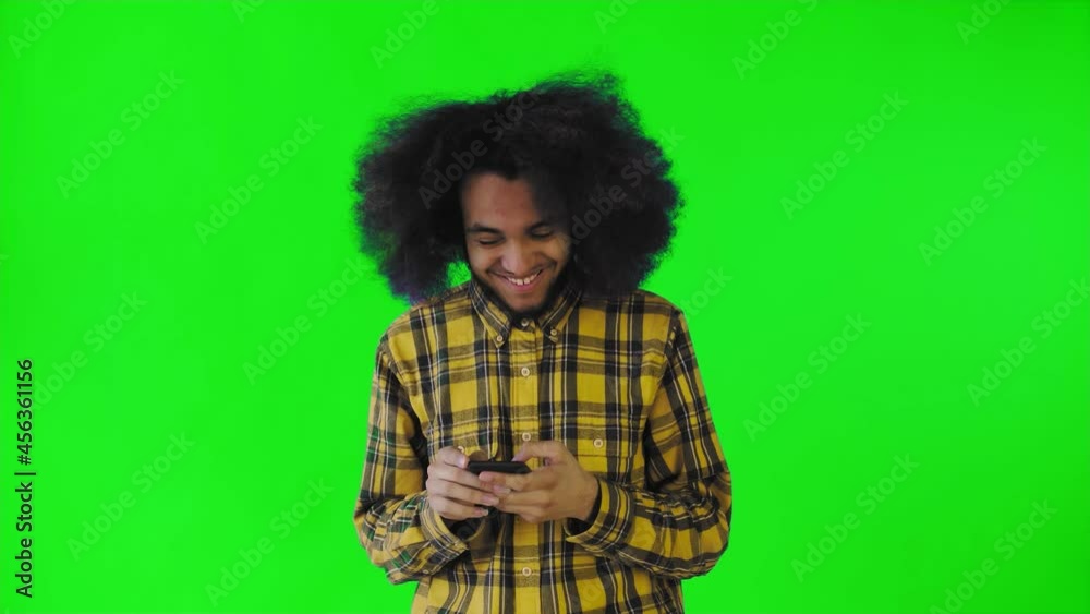 A young man with an African hairstyle on a green background looks at the phone and happily texts with someone. Emotions on a colored background A young man with an African hairstyle on a green background looks at the phone and happily texts with someone. Emotions on a colored background