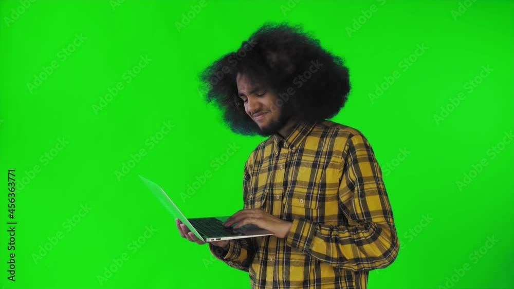 A young man with an African hairstyle on a green background is typing on a laptop. On a colored background A young man with an African hairstyle on a green background is typing on a laptop. On a colored background