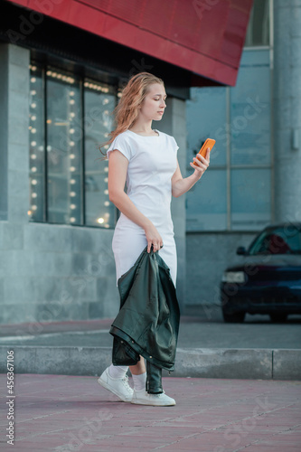 A girl in a plain white dress holds a jacket and a phone against the background of a gray building.