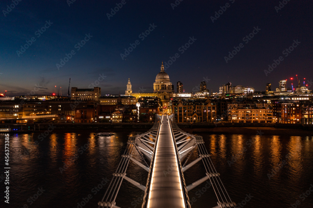 Aerial view of the Millennium Bridge walkway in London city with lights and people walking at night facing towards the church dome