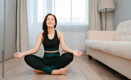 Sporty smiling woman is sitting in the lotus position doing stretching at home in a bright room.