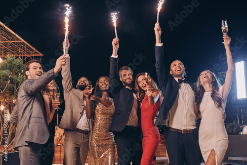 Group of beautiful people in formalwear holding sparklers and smiling while spending time on outdoor party