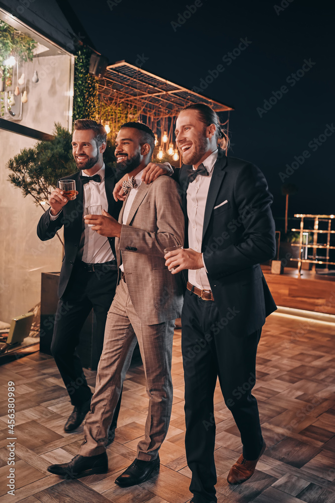 Full length of three handsome men in suits and bowties drinking whiskey and communicating