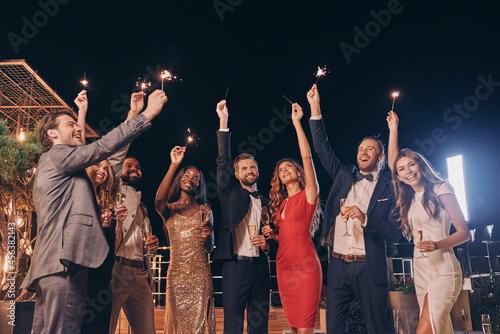 Group of beautiful people in formalwear holding sparklers and smiling while spending time on outdoor party