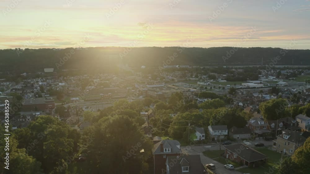A slow orbiting aerial establishing shot of a Pennsylvanian small town at sunset. Pittsburgh suburbs. Bridge over the Ohio River in the distance, high school building in the foreground.  	