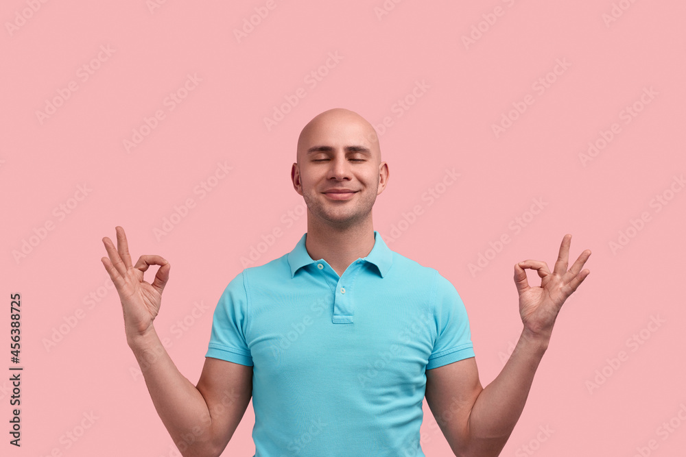 Peaceful pleased bald homosexual man with bristle is practicing yoga, relaxing, makes zen gesture, meditates indoors , closes eyes, gay friendly, wears blue polo shirt, poses over pink background. Peaceful pleased bald homosexual man with bristle is practicing yoga, relaxing, makes zen gesture, meditates indoors , closes eyes, gay friendly, wears blue polo shirt, poses over pink background.