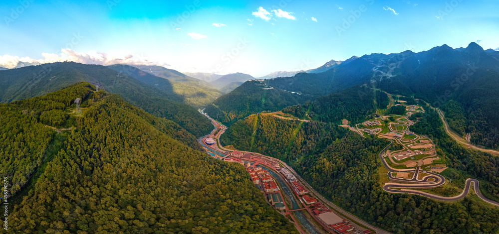 a large aerial panorama of the Mzymta river valley and the hotels of the Rosa Khutor resort, the Sanki luge and bobsleigh complex in the Caucasus mountains on a sunny summer day