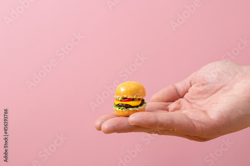 Miniature burger in hand on a pastel pink background. Minimalistic food concept.