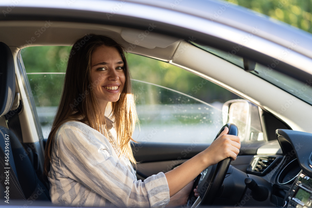 Happy young girl sitting on driver seat in new car joyful smiling hold hands on wheel. Cheerful female driving vehicle looking through open window. Successful woman car owner or getting driver license