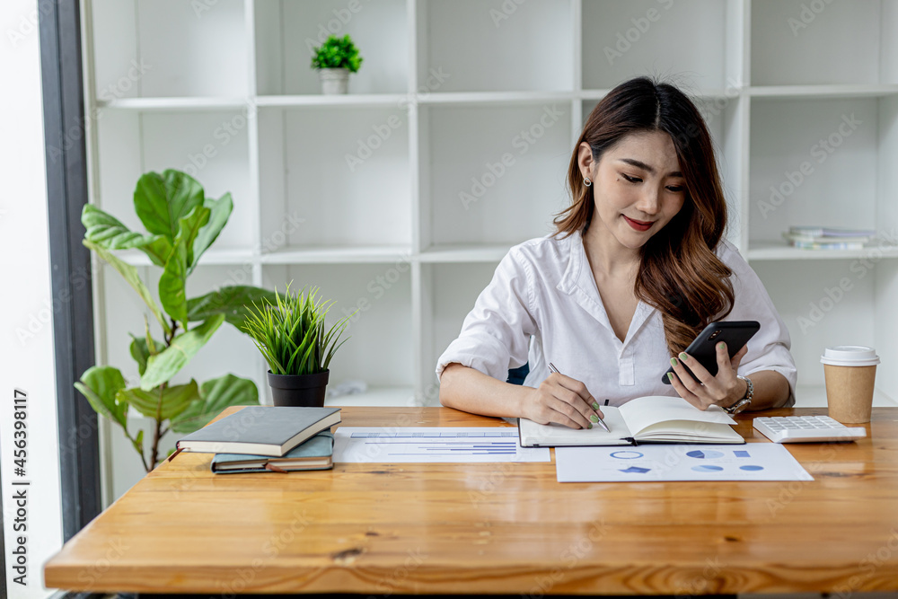 Beautiful Asian woman using a smartphone, businesswoman searching internet on smartphone and taking notes in a notebook, she is working in her room. Concept of Internet search and use of technology. Beautiful Asian woman using a smartphone, businesswoman searching internet on smartphone and taking notes in a notebook, she is working in her room. Concept of Internet search and use of technology.